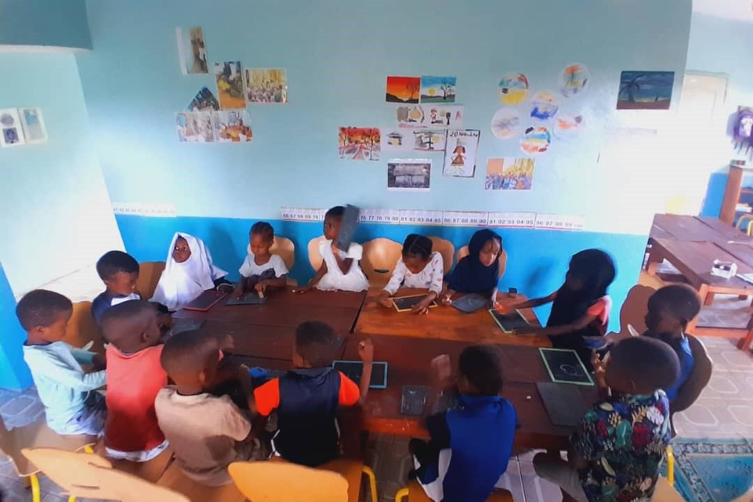 Des élèves installés autour de tables dans leur classe, travaillant sur des ardoises, à la Maison des Enfants Lumières